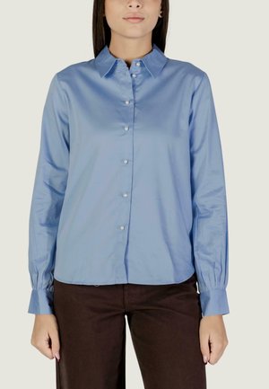 Donna che indossa una camicia azzurro chiaro con bottoni, colletto e maniche lunghe, abbinata a pantaloni marrone scuro, in piedi davanti a uno sfondo neutro.