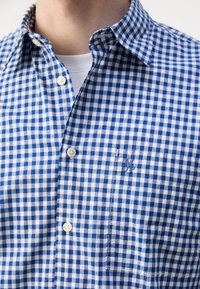 Homme portant une chemise à carreaux bleue et blanche boutonnée par-dessus un T-shirt blanc, montrant le col de la chemise et une poche poitrine avec un petit logo brodé.