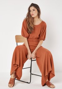 Roestkleurige, wijdvallende jumpsuit met opgezette mouwen en een elastische tailleband. Gladde stof en losse pasvorm. Gecombineerd met bruine slippers.