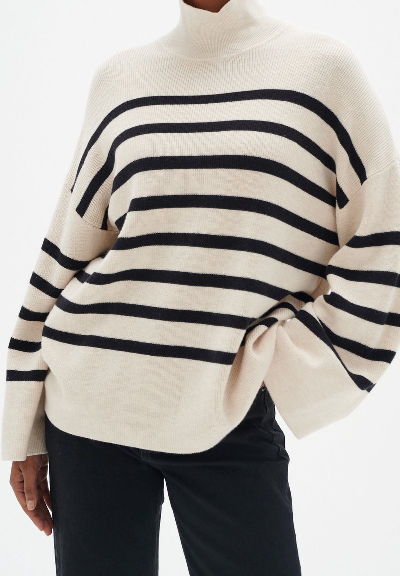 Beige polotröja med svarta horisontella ränder, med avslappnad passform och ribbad textur. Bärs med svarta jeans.