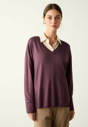 Kvinde med brunt hår iført lilla V-udskåret sweater over beige skjorte med krave og lysebrune bukser, stående mod en ensfarvet lys baggrund.