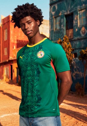 Νεαρός άνδρας στέκεται σε εξωτερικό χώρο φορώντας πράσινη φανέλα ποδοσφαίρου της Σενεγάλης με κίτρινο περίγραμμα και γεωμετρικά σχέδια, με παλιά κτίρια στο φόντο.
