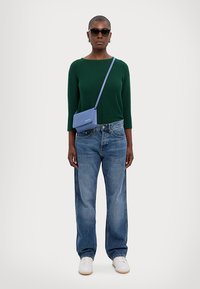 Green knit sweater, blue crossbody bag, blue denim jeans, white sneakers, black sunglasses, standing against a plain background.
