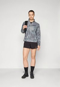 Szary pulower w wzory z półzamkiem, zestawiony z czarnymi szortami. Model trzyma czarny plecak, ma na sobie czarne buty i skarpetki.