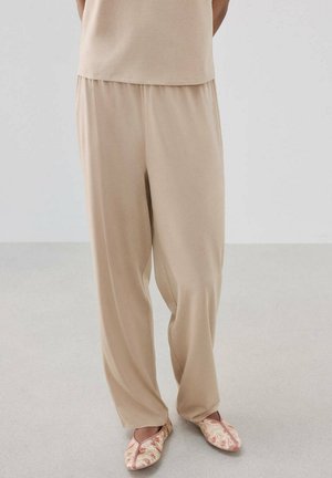 Pantalon beige ample porté avec un haut assorti et des chaussures slip-on rose clair à motifs, sur une personne debout sur un sol clair.