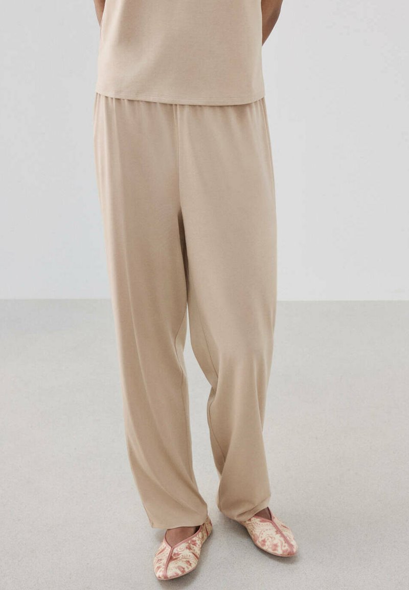 Pantalon beige ample porté avec un haut assorti et des chaussures slip-on rose clair à motifs, sur une personne debout sur un sol clair.