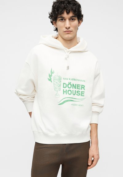 Νέος άνδρας με σγουρά μαλλιά που φοράει λευκό φούτερ με κουκούλα με πράσινο κείμενο «Döner House» και γραφικό σουβλάκι ντονέρ, συνδυασμένο με καφέ παντελόνι.