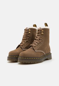 Botas de ante marrón con cuello de piel sintética, ojales de metal y suela de goma negra. Costuras amarillas acentúan el diseño. Estilo con cordones.