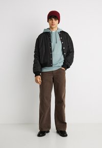 Νέος άνδρας που στέκεται με τα χέρια στις τσέπες, φορώντας μπορντό σκουφί, γαλάζιο φούτερ με κουκούλα, μαύρο μπουφάν bomber, καφέ παντελόνι και μαύρα παπούτσια.