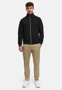 Svart zip-up-jacka med hög krage och vit dragkedja, bärs med beige byxor och vita sneakers. Tyget verkar slätt, med en figursydd design.