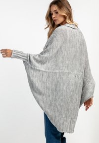 Szary sweter w stylu poncho, luźny krój, szerokie rękawy, ściągacze i wzór teksturowy. Charakteryzuje się lekkim opadaniem i zaokrąglonym dołem.
