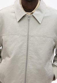 Chaqueta gris claro con cremallera y cuello clásico, con forro interior acolchado y puños acanalados. Textura suave, diseño minimalista.