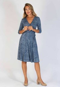 Niebieska sukienka w wzory z rękawami o długości 3/4, wcięciem w talii i spódnicą do kolan. Założona z beżowymi butami na blokowym obcasie.