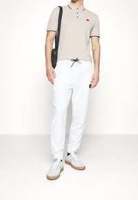 Beige polo shirt met zwarte ribgebreide kraag en een rood logo; witte trainingbroek met een zwarte koord; grijze sneakers met gumzolen.
