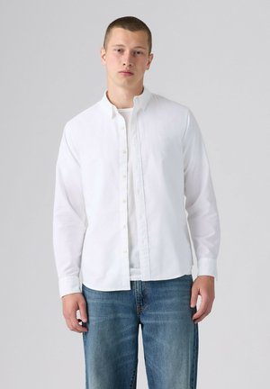 Jeune homme portant une chemise blanche boutonnée sur un t-shirt blanc et un jean bleu, debout devant un fond uni gris clair.