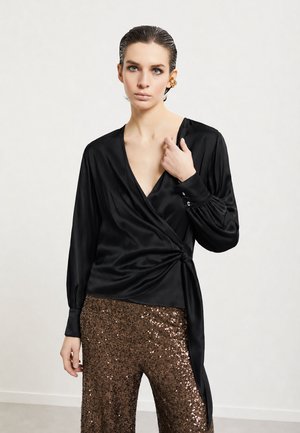 Vrouw met naar achter geslagen haar, die een zwarte satijnen wikkelblouse en glanzende bruine paillettenbroek draagt, staat tegen een lichte achtergrond.