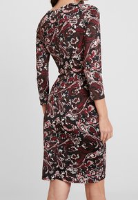 Kvinna med en figurnära knälång klänning med långa ärmar och ett paisley-mönster i röd, vit och svart, sedd bakifrån.