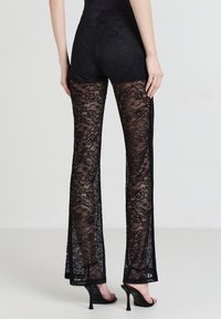 Pantaloni neri in pizzo con un design trasparente, caratterizzati da motivi floreali e una vita nera aderente. Le gambe svasate e i lati aperti esaltano il look.