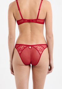 Rotes Dessous-Set aus transparenter Mesh und floralem Spitze. Der BH hat verstellbare Träger und einen Hakenverschluss; der Tanga verfügt über ein Schlüsselloch-Detail.
