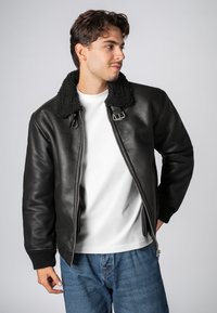 Zwarte leren bomberjack met een schapenvachten kraag, ritssluiting, geribbelde manchetten en een gladde textuur. Draag het over een wit shirt en jeans.