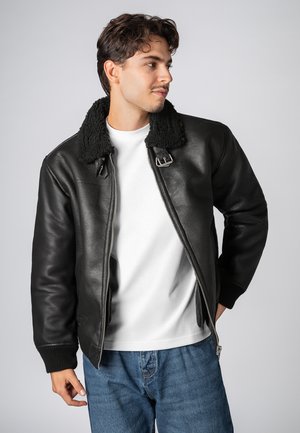 Zwarte leren bomberjack met een schapenvachten kraag, ritssluiting, geribbelde manchetten en een gladde textuur. Draag het over een wit shirt en jeans.