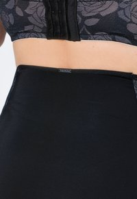 Schwarze, hochtaillierte Hose aus glattem Material mit dezentem Glanz, ausgestattet mit einem kleinen Markenetikett am Bund und einer Spitzen-Mesh-Detail darüber.