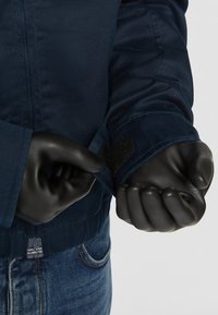 Marineblå jakke med en glatt tekstur, med borrelåser på ermene og elastikk i midjen. Hender i svarte hansker justerer ermekanten.