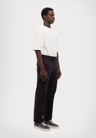 Camiseta blanca de algodón, corte relajado, pantalones de algodón azul oscuro, zapatillas grises con cordones blancos, en una postura de pie frente a un fondo liso.