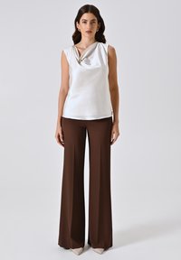 Top senza maniche in raso bianco con dettaglio annodato e accento dorato, abbinato a pantaloni a vita alta in tonalità marrone e a gamba ampia. Scarpe neutre a punta.