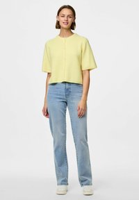 Dzeltena īsā adīta džemperis ar īsām piedurknēm un pogu aizdari, apvienota ar gaiši ziliem taisnajiem džinsiem un baltām kedām.