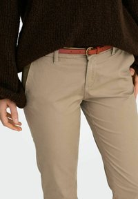 Inimene kannab beežpükse, millel on õhuke punane vöö ja paksem tumepruun kampsun, käed lõdvestunult kõrval.