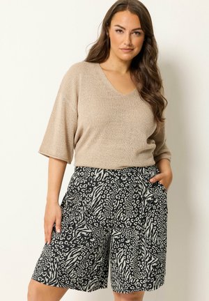 Frau mit langen braunen Haaren, die ein beiges kurzärmeliges Strickoberteil und knielange Shorts mit schwarz-weißem Tiermuster trägt, steht mit einer Hand in der Tasche.