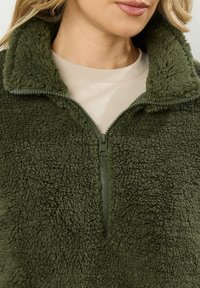 Olijfgroene fleece pullover met een hoge kraag en halve rits. Zachte, gestructureerde oppervlakte met een gladde ritsdetail en een crèmekleurige onderlaag.