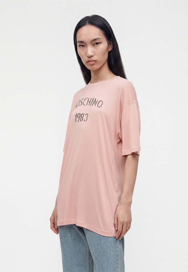Woman with long dark hair wearing a loose pink Moschino 1983 t-shirt and light blue jeans standing against a plain white background.