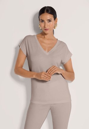 Woman with dark hair in low ponytail wearing beige V-neck short-sleeve top and matching pants, posing with hands clasped.