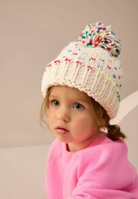 Knitted cream beanie with multicolored pom-pom, featuring bright speckles of pink, blue, and yellow accents; worn with pink sweatshirt.
