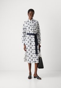 Abito bianco con motivo geometrico blu, maniche lunghe, chiusura con bottoni frontale, cintura legata e spacchi laterali. Scarpe slide nere e borsa tote.