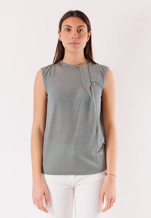 Donna con capelli lunghi e scuri che indossa una canotta grigia senza maniche con piega frontale e pantaloni bianchi, in piedi davanti a uno sfondo chiaro e uniforme.