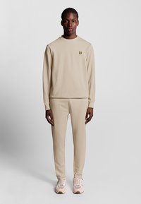 Completo tuta beige con maglia a maniche lunghe e scollo rotondo e pantaloni abbinati, con un piccolo logo giallo sul petto. Tessuto dalla texture liscia.