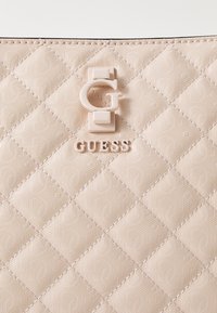 Beige, steppelt szintetikus kézi táska, amely dombornyomott logómintával, fényes G hardver díszítéssel és kiemelkedő név részlettel rendelkezik.
