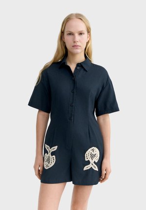 Kobieta ubrana w granatowy kombinezon na guziki z krótkim rękawem i beżowymi koronkowymi akcentami w kształcie ryb na przedniej części spodenek.