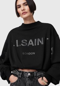 Crni oversized dukserica s natpisom "ALLSAINTS LONDON" u reflektirajućem pismu, u kombinaciji s sivo visokim trapericama.