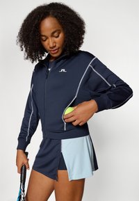 Marinblå idrottsjacka med silverfärgade detaljer och en zilvrig ficka, som håller en tennisboll, i kombination med en tvåfärgad kjol med blått inslag.
