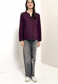 Bordeauxrotes Langarmoberteil mit V-Ausschnitt, kombiniert mit ausgewaschenen grauen Jeans mit geradem Bein und weißen Turnschuhen mit grünen Akzenten.