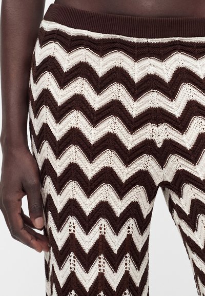 Κοντινό πλάνο σκούρου χρώματος δέρματος χεριού και μέσης που φορά καφέ και λευκό πλεκτό παντελόνι με σχέδιο chevron και ριμπ ζώνη μέσης.