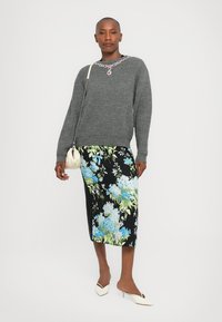 Grauer Strickpullover mit einem besetzten Ausschnitt, kombiniert mit einem schwarzen floral gemusterten Midikleid mit blauen und grünen Mustern. Weiße Absätze und eine kleine Tasche.