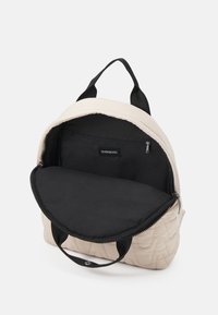 Sac à dos matelassé beige clair avec intérieur noir, fermeture éclair et petite poche intérieure. Deux bretelles noires pour le port.