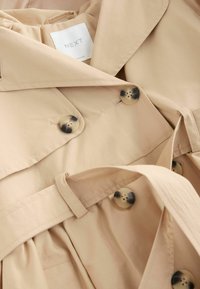 Trench beige con cinturón, grandes botones crema y marrón, y una textura suave. Etiqueta visible en el interior, que indica la marca Next.