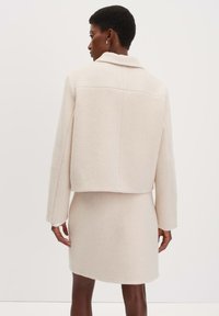 Licht beige cropped jack met lange mouwen en een kraag, gecombineerd met een bijpassende rok tot de knie, beide van een zachte, textuurvolle stof.