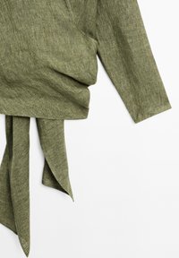 Haut à manches longues en tissu vert olive texturé avec un détail de lien à nouer étendu suspendu sur fond blanc.